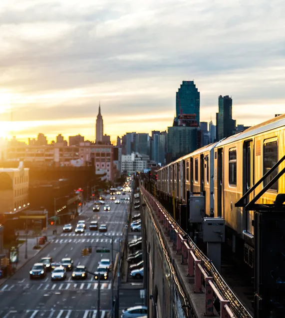 A high-angle view of an elevated train running beside a busy city street at sunset. Golden light glows on the train as cars below create blurred streaks with their headlights. The Manhattan skyline, including the Empire State Building, is silhouetted against a warm orange-to-blue sky.