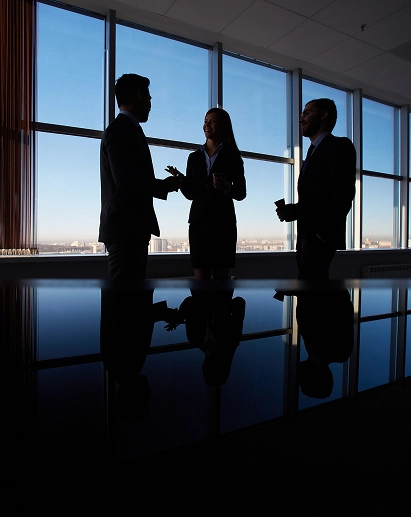 Three silhouetted professionals, two men and one woman, stand talking in a bright office