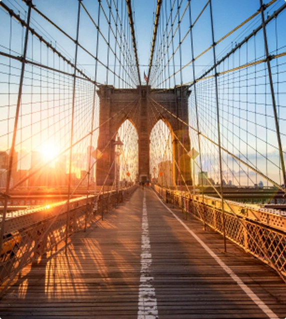 Brooklyn Bridge pedestrian walkway at sunrise, with sunlight streaming from the left, casting long shadows across the wooden planks toward the iconic stone towers and suspension cables