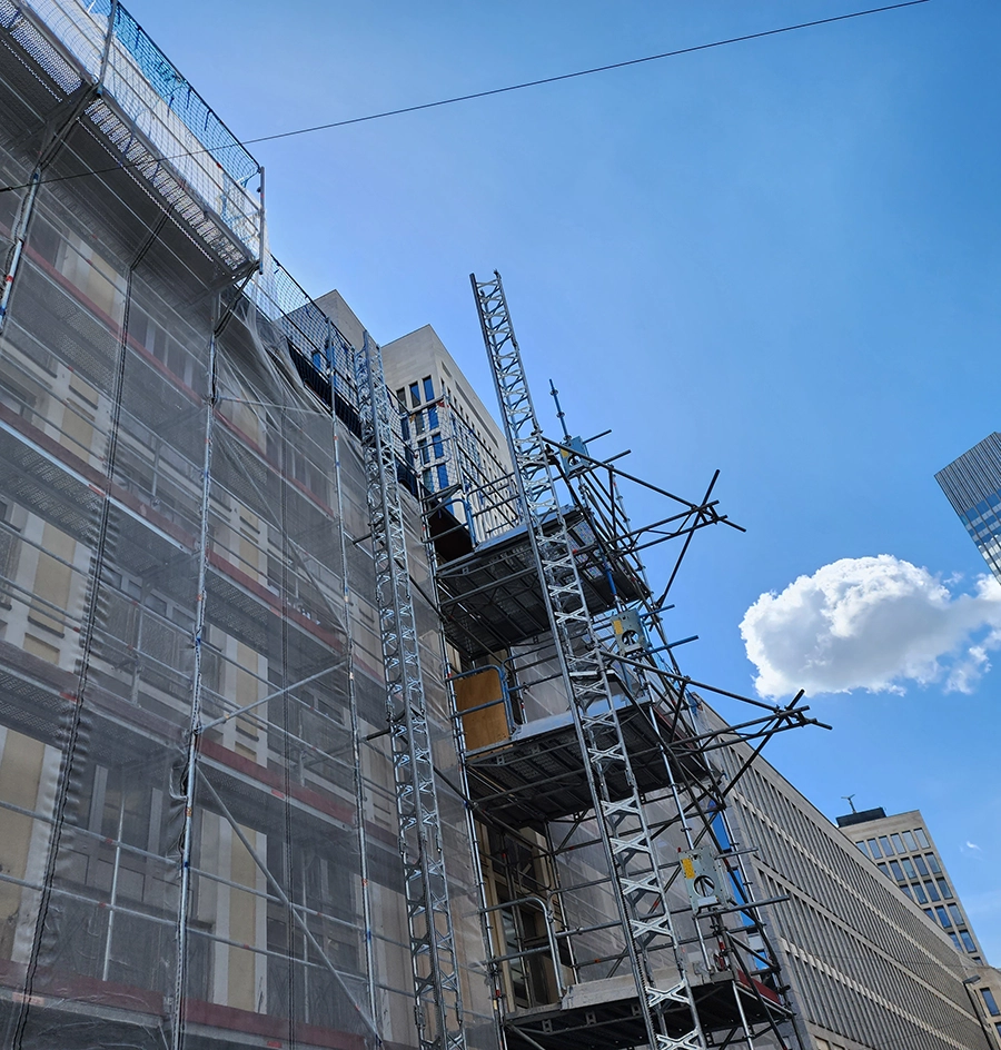 Low-angle view of a large building wrapped in scaffolding and light gray netting under a bright blue sky with a single white cloud.