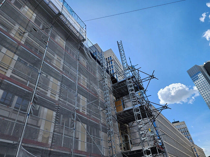 Low-angle view of a large building wrapped in scaffolding and light gray netting under a bright blue sky with a single white cloud.